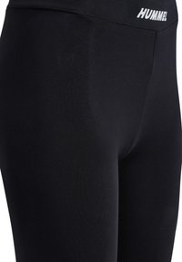 Zwarte leggings gemaakt van rekbare stof. Heeft een brede tailleband met de merknaam "HUMMEL" in witte letters aan de voorkant.