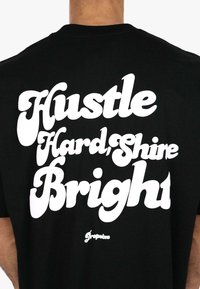 Sort t-shirt med stort hvidt, stiliseret tekst, der står "Hustle Hard, Shine Bright," med "Gropetize" logoet nederst.