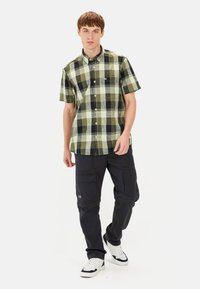 Korte mouw geruit overhemd in groen en zwart, met een button-down kraag en een logo op de borst, gecombineerd met marineblauwe cargobroek en witte sneakers.