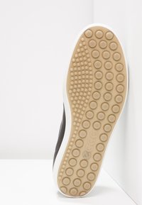 Semelle de chaussure noire avec une surface en caoutchouc texturée présentant des motifs circulaires, encadrée par une couche blanche, sur un fond clair.