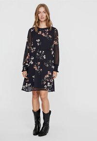 Robe florale noire avec de longues manches transparentes, taille élastique et jupe fluide. Présente des motifs floraux roses et blancs sur l'ensemble.