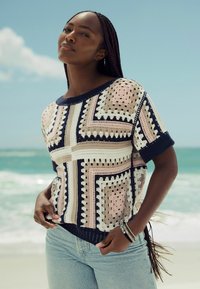 Häkelpullover mit geometrischen Mustern in Creme, Rosa und Marineblau. Kurze Ärmel, lockere Passform und strukturierte Oberfläche. Kombiniert mit blauen Jeans.