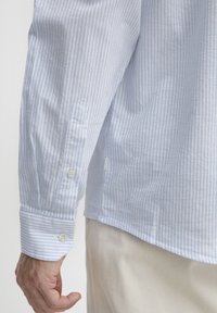 Manga larga a rayas azules y blancas con puño sobre el brazo de una persona que lleva puesta un pantalón color crema.