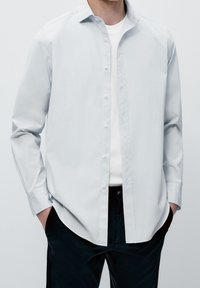 Camicia azzurra chiaro in cotone morbido, con colletto classico e maniche lunghe, indossata sopra una t-shirt bianca, abbinata a pantaloni scuri.