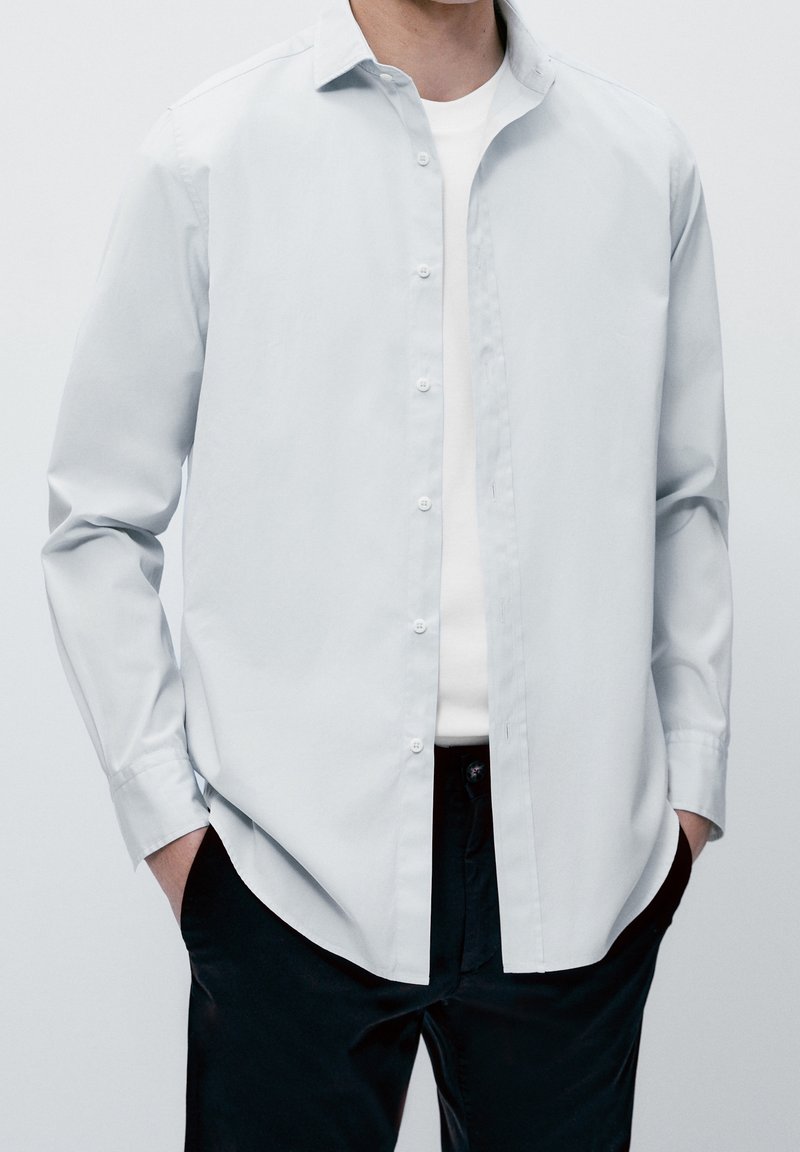 Camicia azzurra chiaro in cotone morbido, con colletto classico e maniche lunghe, indossata sopra una t-shirt bianca, abbinata a pantaloni scuri.