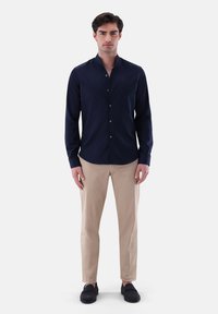 Camicia blu navy a maniche lunghe con colletto alla coreana, chiusura con bottoni e texture liscia, abbinata a pantaloni beige e scarpe slip-on scure.