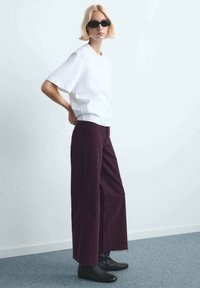 Oseba s kratkimi blond lasmi, ki nosi črna sončna očala, belo srajco s kratkimi rokavi, široke temno vijolične hlače in črne gležnjarje, stoji bočno.