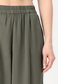 Pantalon vert olive, à taille élastique, avec une texture lisse, comportant des poches latérales et une coupe évasée. Vue en gros plan de la taille et de la main.