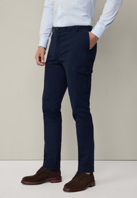 Marineblauwe cargobroek met een gladde textuur, voorzien van zijzakken en een knoopsluiting, gecombineerd met bruine suède enkellaarsjes.