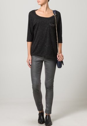 Mujer que lleva un top negro con motas, pantalones grises ajustados, zapatos negros sin cordones y lleva un bolso de hombro azul marino con cadena como tira.