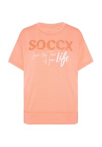 Camiseta rosa de gran tamaño con mangas cortas. Presenta texto naranja brillante "SOCCX" y la frase en cursiva blanca "Disfruta el momento de tu vida". Tejido de mezcla de algodón.