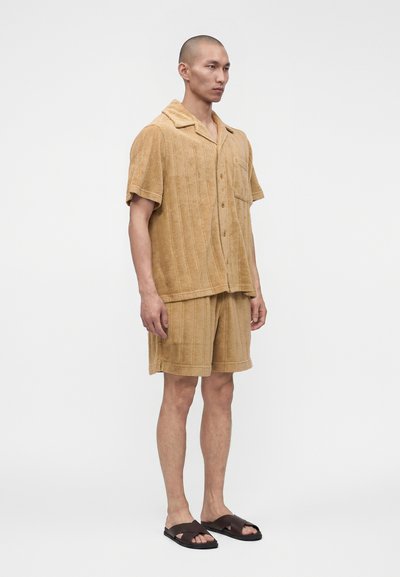 Homme portant une chemise à manches courtes beige texturée boutonnée, un short assorti et des sandales marron foncé, debout sur fond blanc.