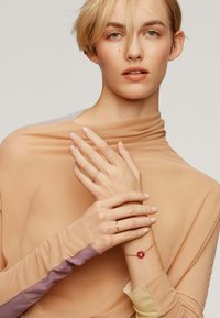 Top ligero y transparente en tono beige, con mangas largas y detalles en púrpura y verde apagado; la modelo lleva anillos finos y una pulsera con un detalle rojo.