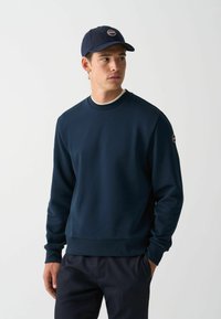 Marineblauwe sweatshirt met geribbelde ronde hals en manchetten, gecombineerd met donkere marineblauwe broeken en een bijpassende pet met een rond logo.
