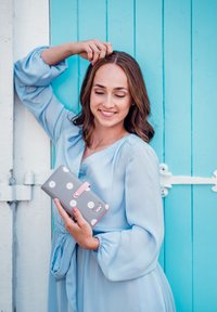 Gray wallet with white polka dots and a pink strap, held against a blue wooden background. Features a smooth texture and rectangular shape.