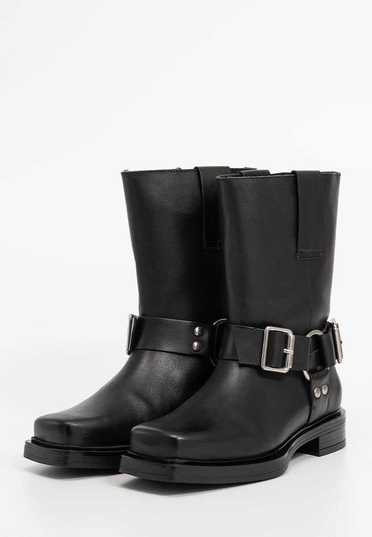 Bottes chevilles en cuir noir avec une semelle plate, dotées d'une sangle à boucle sur le côté et d'accents en métal argenté. Texture lisse avec un design structuré.