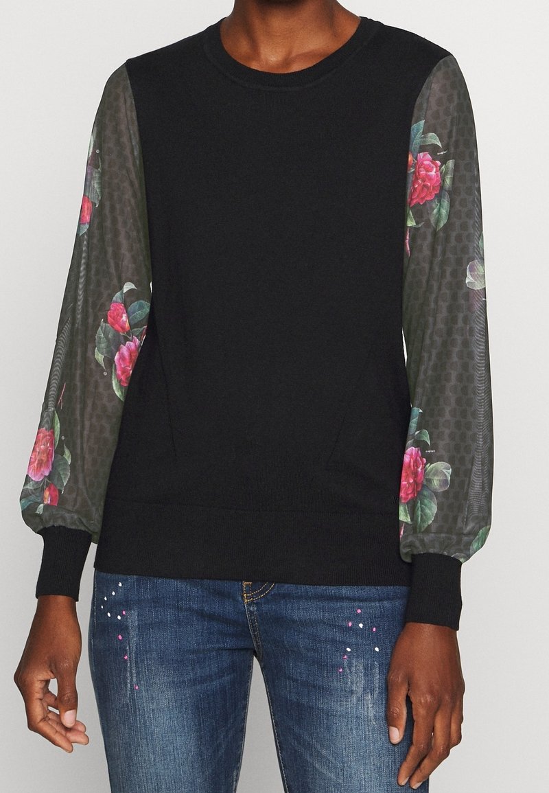 Svart tröja med genomskinliga ballongärmar prydda med rosa blommönster, bärs tillsammans med blå jeans som har vit och rosa färgspillet detaljer.