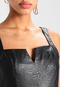 Gilet en cuir noir avec un décolleté carré et de fines bretelles, présentant une texture subtile et un détail central en encoches.