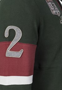 Tummanvihreä kangaspulloveri, jossa viininpunainen raita, suuri harmaa kirjailtu numero '2' sekä valkoinen yksityiskohta helmassa.