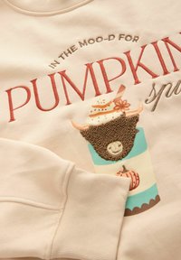 Sweatshirt beige clair avec des cuffs côtelés, orné d'une broderie colorée représentant une tasse, un gâteau et une tête de taureau avec un visage souriant.