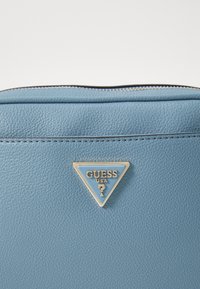 Borsa in pelle strutturata di colore azzurro chiaro con una placca logo triangolare in tonalità oro con dettagli blu, sopra una chiusura a zip.