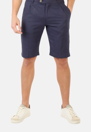 Uomo che indossa pantaloncini blu navy su misura e sneaker bianche, in piedi con le mani in tasca su uno sfondo semplice.