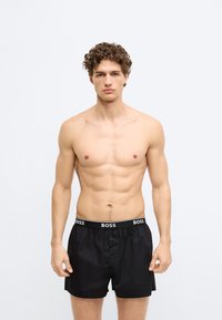 Čierne boxerky s elastickým pásom označeným "BOSS." Ľahký materiál, voľný strih a zapínanie na gombík vpredu.