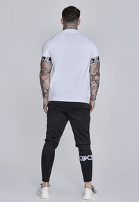 Hvid tætsiddende t-shirt med sorte logo-striber på ærmerne, parret med sorte tapered joggers med logo-detalje og hvide sneakers.