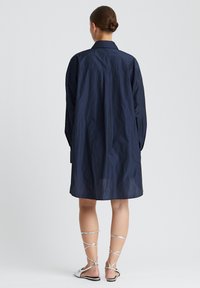 Marineblaues, übergroßes Hemdkleid mit langen Ärmel, Kragen und gekräuselter Textur, das knapp über den Knien endet. Getragen mit silbernen Riemchensandalen.