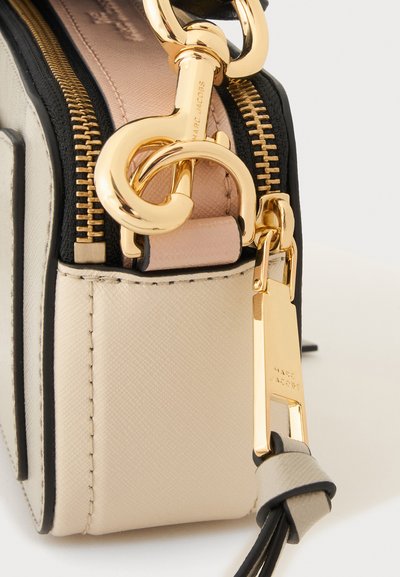 Γωνία μπεζ και μαύρης τσάντας Marc Jacobs με χρυσό κούμπωμα, φερμουάρ και επώνυμες λεπτομέρειες μεταλλικών εξαρτημάτων.