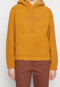 Fleece huvtröja i senapsgul med kenguruficka, dragskohuva och "OBEY"-logotyp i mörkbrunt över bröstet.