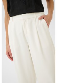 Bílé široké kalhoty s plisováním, s předními kapsami a hladkou texturovanou látkou. Kombinované s černým topem, ruka v kapse.