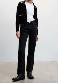 Cardigan noir texturé avec des bordures blanches contrastantes, porté sur une chemise blanche, associé à un jean noir taille haute et des bottines.