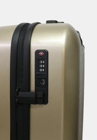 Maleta de carcasa dura en dorado metálico con acabado texturizado, que presenta un zipper negro, cerradura de combinación y doble cierre de seguridad.