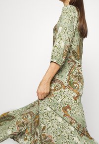 Dlouhé zelené šaty s paisley a květinovými vzory, s tříčtvrtečními rukávy a splývavou sukní. Měkká textura látky, volný střih.