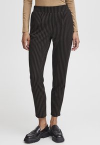 Pantaloni neri a righe con accenti dorati verticali, slim fit, vita con pieghe, abbinati a mocassini neri spessi.
