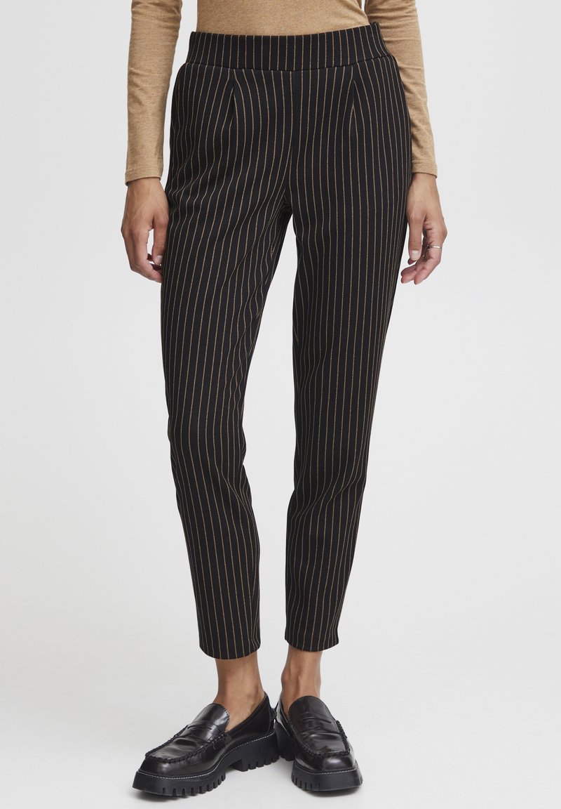 Pantaloni neri a righe con accenti dorati verticali, slim fit, vita con pieghe, abbinati a mocassini neri spessi.