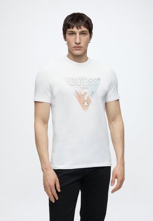 Homme portant un t-shirt blanc Guess avec un logo triangulaire multicolore et un pantalon noir, debout sur fond blanc.