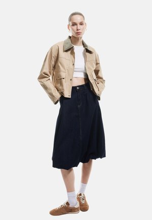 Beige cropped jakke med grøn krave, hvid crop top, mørkeblå A-formet nederdel, hvide sokker og brune sneakers, med synlige sømme.