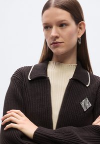 Cardigan marron côtelé avec accents de col blancs, écusson logo et fermeture éclair. Porté sur un pull côtelé crème, avec manches longues.