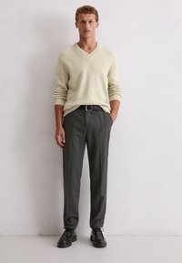 Helle beige V-Ausschnitt-Strickjacke, strukturierter Strick, kombiniert mit maßgeschneiderten dunkelgrauen gestreiften Hosen. Zubehör umfasst einen schwarzen Gürtel und schwarze Schuhe.