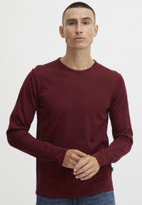 Langermet burgunder skjorte laget av mykt stoff, med rund halslinning og enkelt design. Båret med blå jeans, hender samlet.