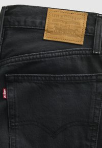 Jean en denim noir avec une poche arrière visible, un patch en cuir marron Levi Strauss & Co et une petite étiquette en tissu rouge Levi's sur la couture latérale.