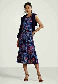 Cvetlična obleka v modri barvi, s rdečimi in rumenimi poudarki. Oprijet zgornji del in razširjen krilo, v kombinaciji s temnim pletenim kardiganom. Rjave sanjske sandale.