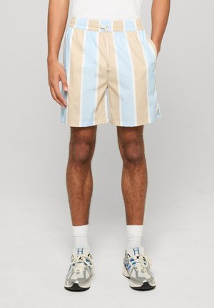 Mand iført beige og lyseblå lodrette stribede shorts, hvide sokker og grå sneakers, stående med hænderne i lommerne.