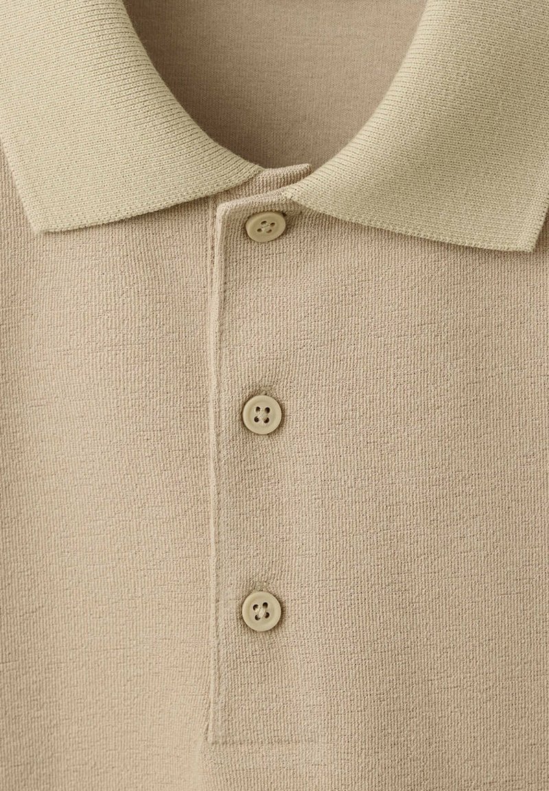 Polo de punto beige claro con tres botones a juego y un cuello plano, mostrando la textura de la tela en primer plano.