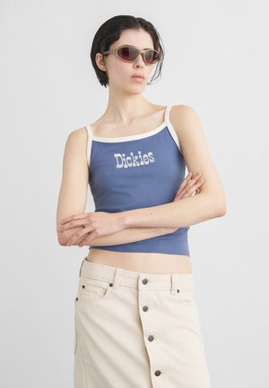 Lühikeste tumedate juustega naine, kandes päikeseprille, sinist tankitoppi, millel on kiri "Dickies", ja beeži nööpidega seelikut, käed rüpes.