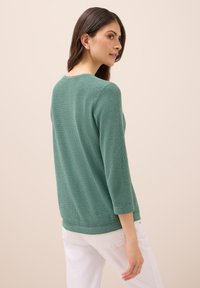 Grüner gestrickter Pullover mit rundem Ausschnitt und drei Viertelärmeln. Verfügt über ein strukturiertes Muster und eine lockere Passform. Kombiniert mit weißen Hosen.