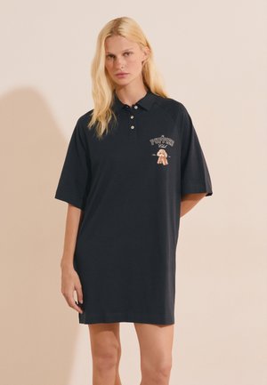 Mujer rubia que lleva un vestido polo negro holgado con el logo "Puppies Club" y un pequeño gráfico de un perro en el pecho izquierdo, de pie sobre un fondo liso.