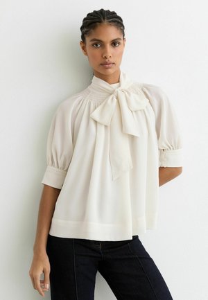 Crèmekleurige blouse met een geknoopte kraag, opgepofte mouwen en een gerimpeld inzetstuk. Gladde textuur en lichtgewicht stof, gecombineerd met donkere spijkerbroek.